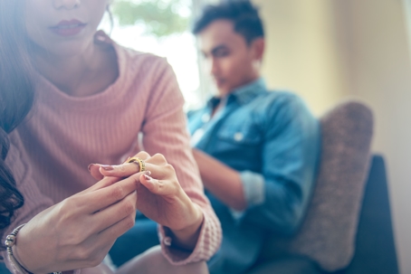 Close-up of a Sad Wife want to Divorce Lamenting Holding the Wedding Ring with Blurred Man on Sofa Background.